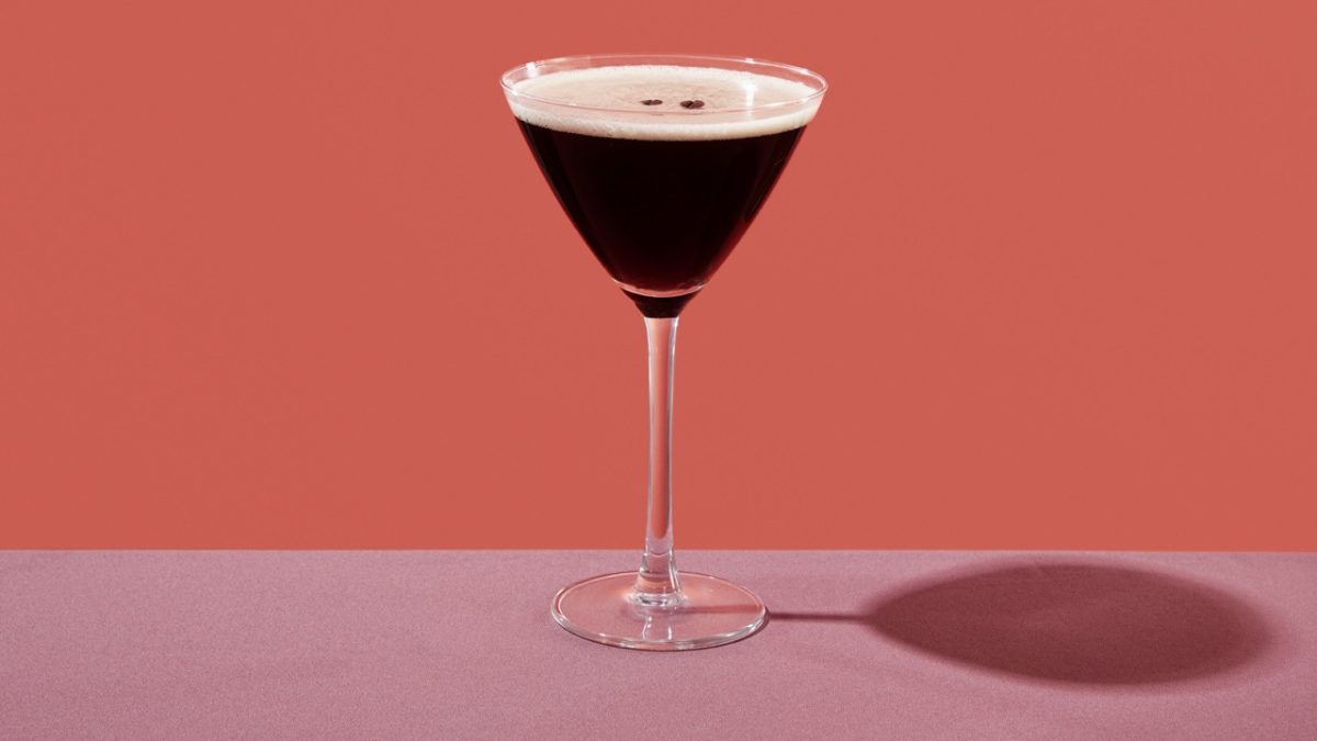an espresso martini served in a cocktail glass, on a table set with a maroon tablecloth, against a coral pink background