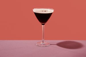 an espresso martini served in a cocktail glass, on a table set with a maroon tablecloth, against a coral pink background